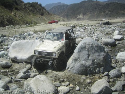 1987 Suzuki Samurai - Suzuki Samurai tackling rough terrain in a rocky riverbed, with other vehicles visible in the distance.