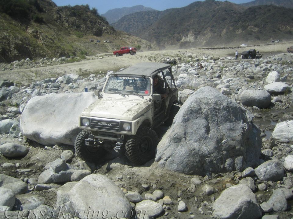 1987 Suzuki Samurai navigating a rocky riverbed, with a red pickup truck in the background