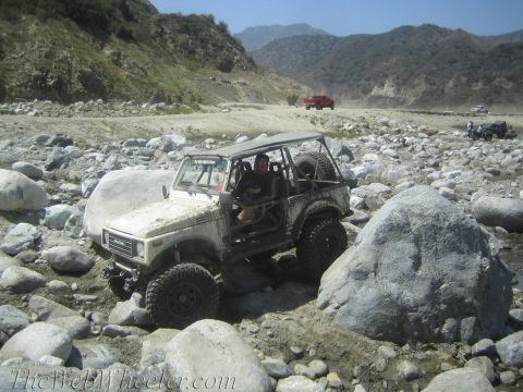 1987 Suzuki Samurai - A white 1987 Suzuki Samurai tackles a challenging rocky terrain in a dry riverbed, with other vehicles visible in the background.