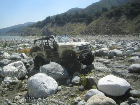 1987 Suzuki Samurai - A white 1987 Suzuki Samurai navigates a rocky terrain near a body of water under a clear sky.