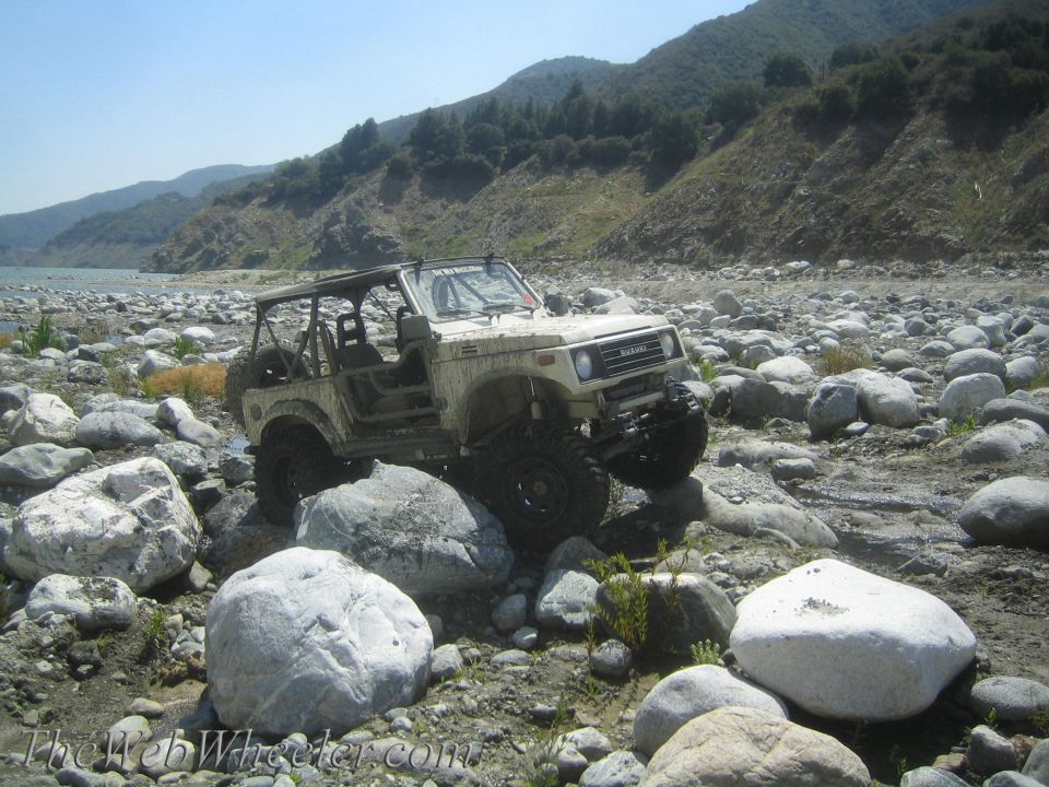 White 1987 Suzuki Samurai off-roading on a rocky riverbed, front-right wheel elevated