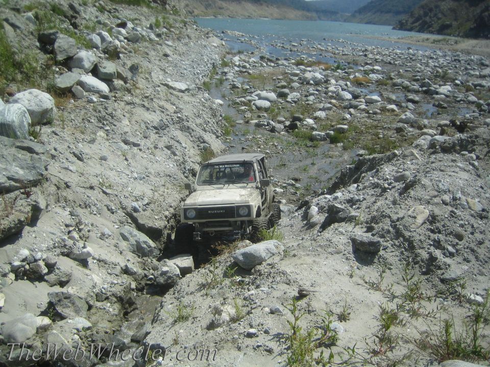 1987 Suzuki Samurai navigating a rocky, dry riverbed with a body of water in the background