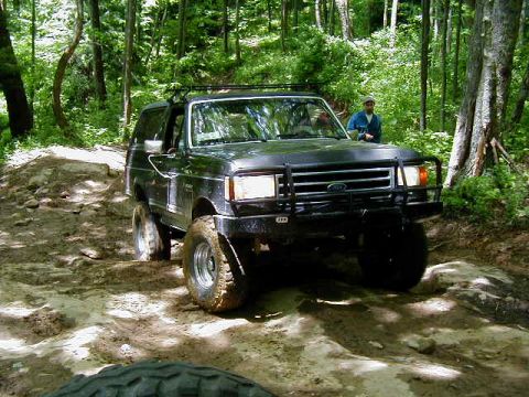 1990 Ford Bronco - A Ford Bronco navigates a challenging off-road path surrounded by trees.