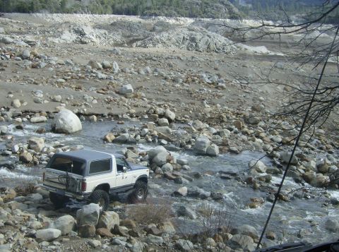 1986 Ford Bronco - A white Ford Bronco navigates a shallow, rocky riverbed with a mountainous, debris-filled landscape in the background.