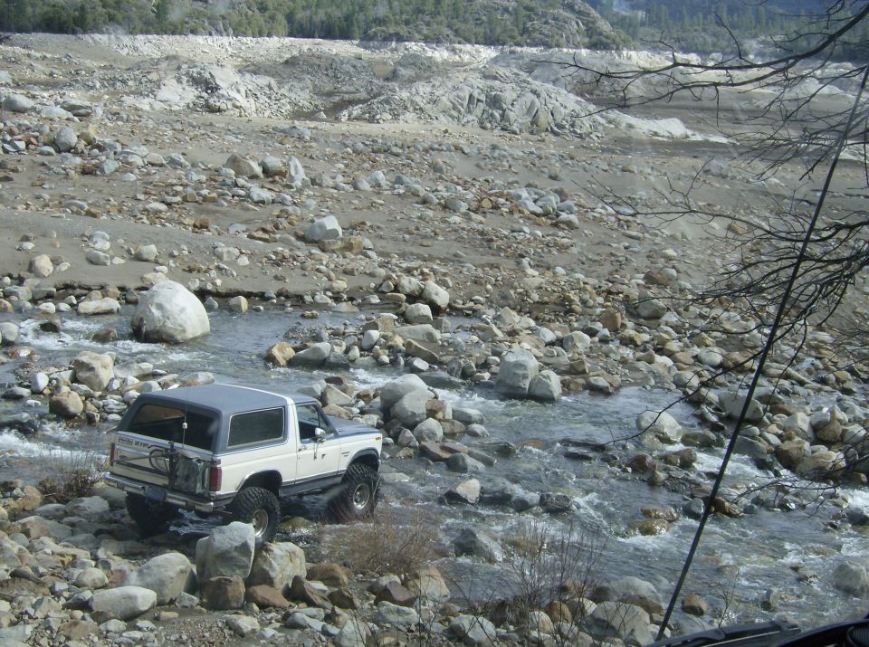 White Ford Bronco with spare tire on back driving in a rocky stream bed