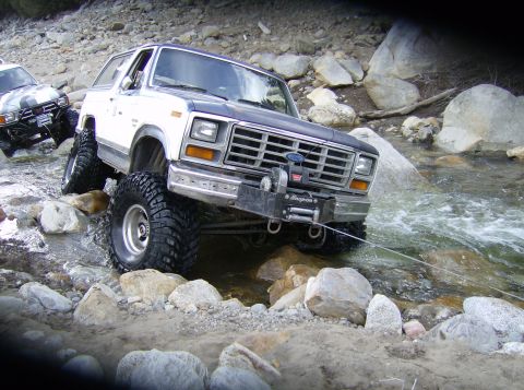 1986 Ford Bronco - A white and blue 1980 Ford Bronco navigates a rocky stream, equipped with a winch and large off-road tires.