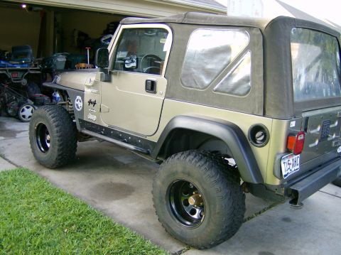2004 Jeep Wrangler - A tan Jeep Wrangler with a soft top and large tires is parked in front of a garage.