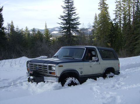 1986 Ford Bronco - White and blue two-tone Ford Bronco buried to the axles in deep snow on a mountain ridge, seen from the driver 3/4 front with a winch line…