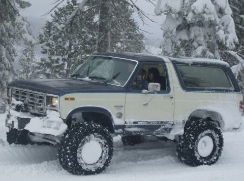 1986 Ford Bronco - A two-tone Ford Bronco navigates a snowy landscape with large off-road tires.