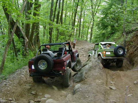 1990 Ford Bronco - Two Ford Broncos navigate a rocky, wooded trail.