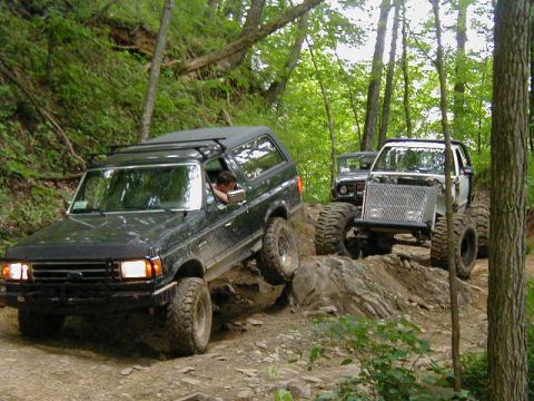 1990 Ford Bronco - Two modified SUVs navigate a challenging off-road trail through a wooded area.