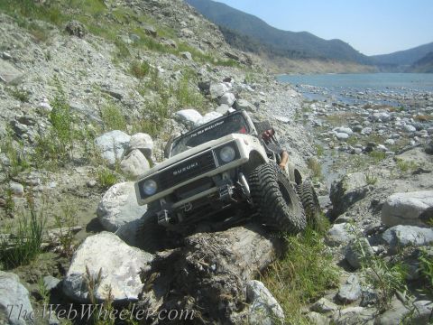 1987 Suzuki Samurai - A white 1987 Suzuki Samurai tackles a challenging off-road course, with a driver visible behind the wheel.