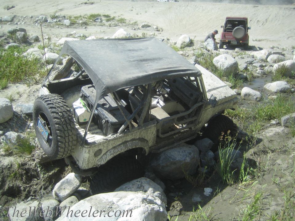 Mud-covered Suzuki Samurai with a soft top, stuck on rocks in a stream. A second red Suzuki Samurai is visible in the background