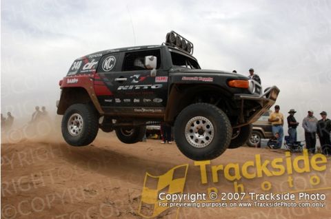 1992 Ford Explorer - Toyota FJ Cruiser airborne during a desert race event, with onlookers and another vehicle visible.