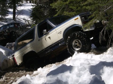 1986 Ford Bronco - A white and blue Ford Bronco navigates a snowy trail, with another vehicle visible in the background.