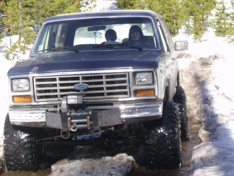 1986 Ford Bronco - A modified Ford Bronco navigates a snowy and muddy trail, its large tires and winch indicating off-road capability.