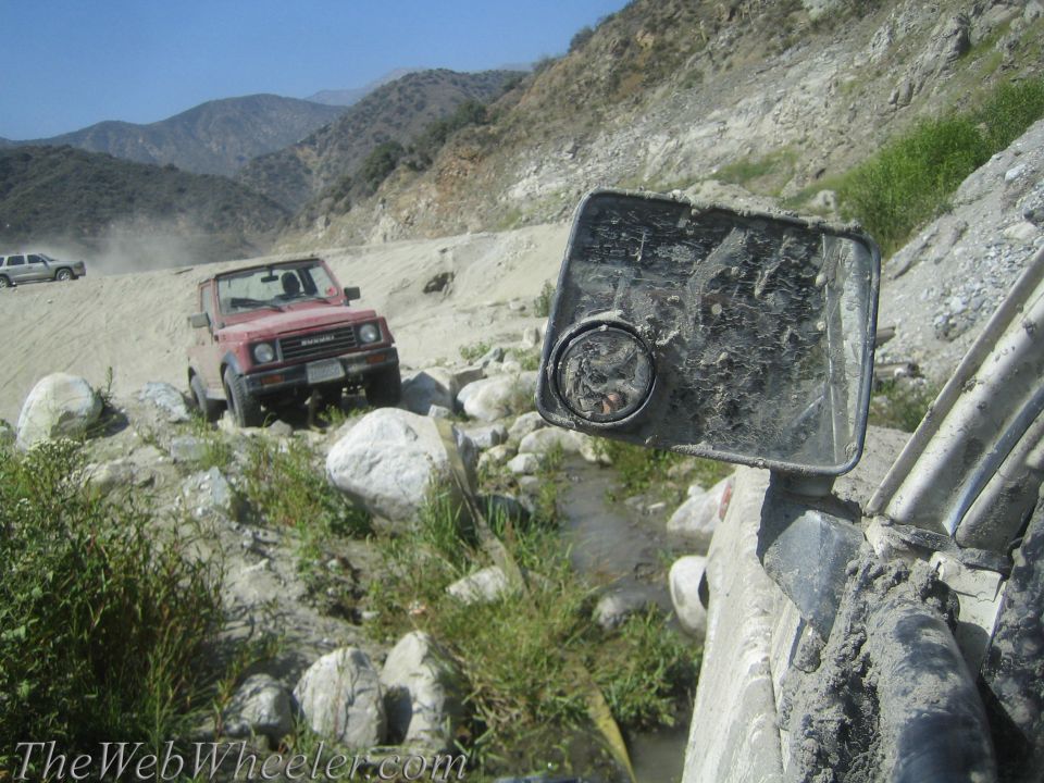 Red Suzuki Samurai driving on a rocky, dusty trail with another vehicle in the background