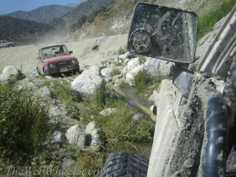 1987 Suzuki Samurai - A red 1987 Suzuki Samurai is seen off-roading on a dusty, rocky trail, with another vehicle visible in the background.