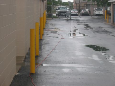 1991 Ford Bronco - A wet asphalt area with yellow bollards and parked vehicles visible through a fence.