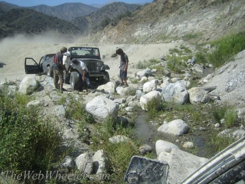 1987 Suzuki Samurai - A group of people work on changing a tire on a Jeep Wrangler in a rocky, off-road environment.