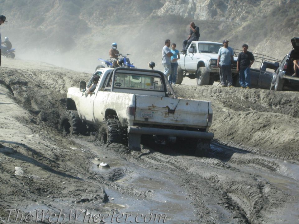 White pickup truck stuck in mud, with people watching and other vehicles nearby