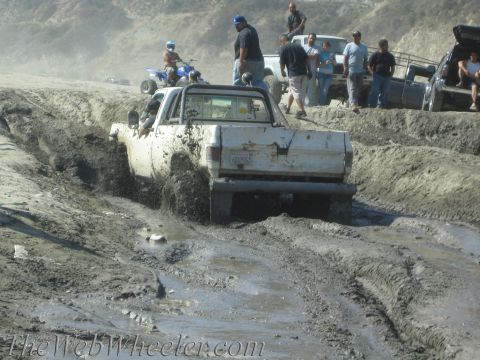 1987 Suzuki Samurai - A white pickup truck struggles through thick mud, with other vehicles and people visible in the background.