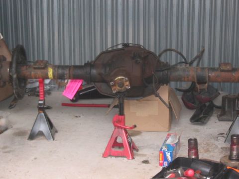 1991 Ford Bronco - A rusty rear axle assembly is supported by jack stands in a garage, with various tools and parts scattered around.