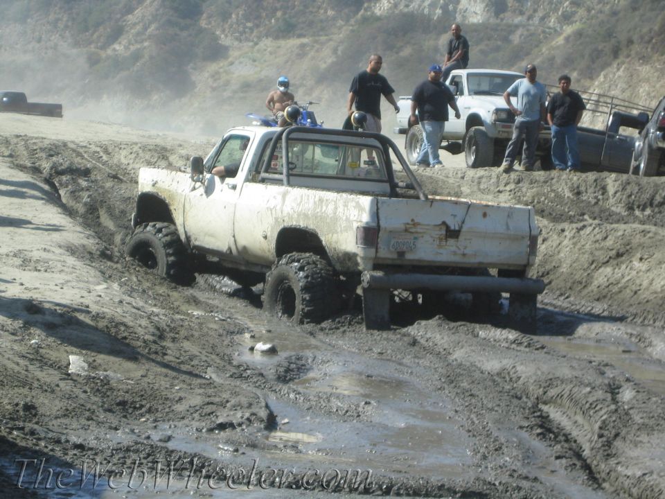White pickup truck with large tires stuck in mud, other vehicles and people in background