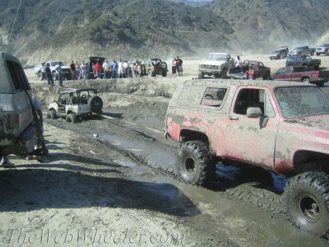 1987 Suzuki Samurai - Several off-road vehicles navigate a challenging muddy terrain under a bright sky.