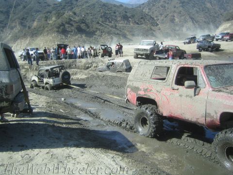 1987 Suzuki Samurai - Off-road vehicles are stuck in the mud during an event, with spectators observing from the bank.