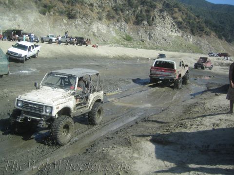 1987 Suzuki Samurai - A white 1987 Suzuki Samurai navigates a muddy off-road course, with a red truck following closely behind.