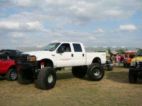 1978 Ford Bronco - A white lifted Ford F-250 Super Duty truck with massive tires is parked at an outdoor event with other vehicles.