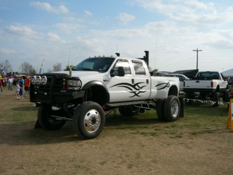 1978 Ford Bronco - A heavily modified white dually pickup truck is parked on a grassy field with other vehicles in the background.
