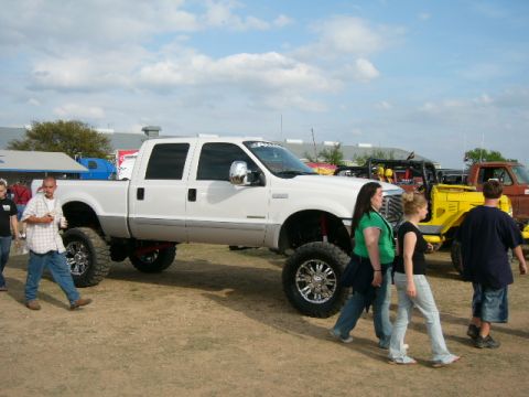 1978 Ford Bronco - A lifted white pickup truck is parked at an outdoor event with other vehicles and people nearby.