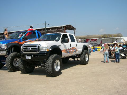 1978 Ford Bronco - A lifted white Ford F-Series truck is parked next to a blue truck at an outdoor event.