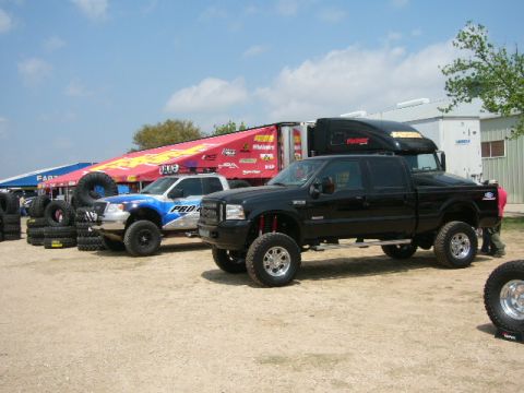 1978 Ford Bronco - Lifted pickup trucks and a semi-trailer are parked outdoors under a partly cloudy sky.