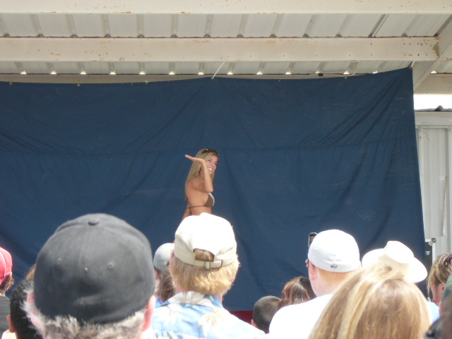 Woman in bikini on stage with a blue backdrop, viewed from behind an audience