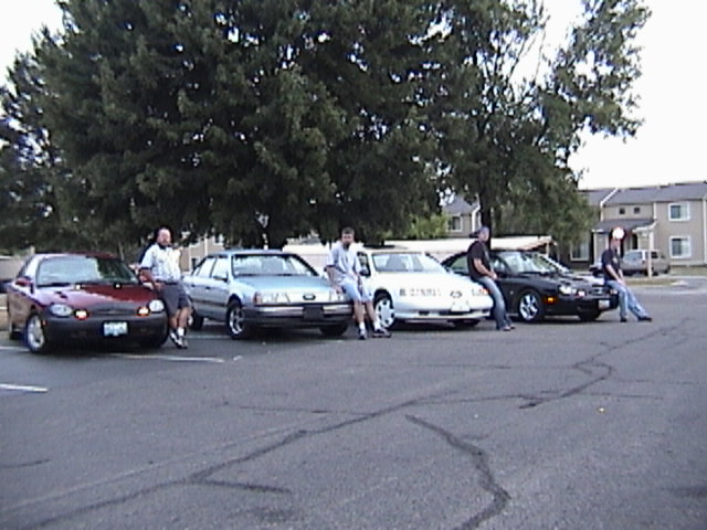 Four sedans parked in a row in a lot with four people standing by them