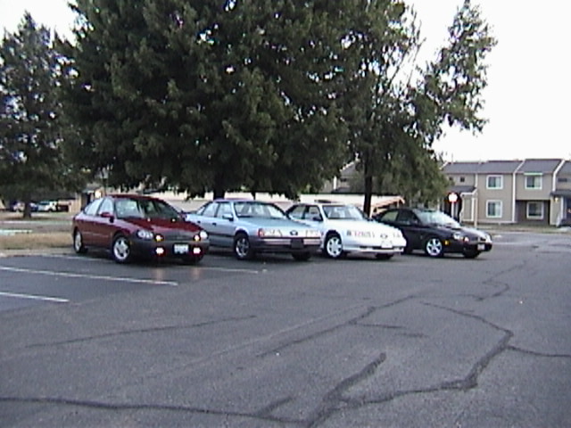 Four sedans parked in a lot with trees and buildings in the background