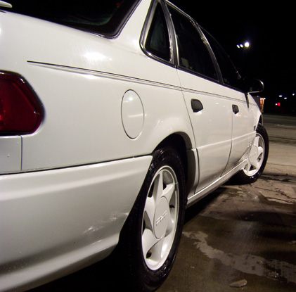 1993 Ford Taurus - A white 1993 Ford Taurus sedan is viewed from the rear quarter panel at night.