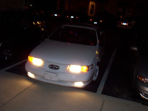 1993 Ford Taurus - Front view of a white 1993 Ford Taurus parked in a lot at night with its headlights illuminated.