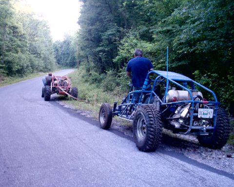 1989 Ford Bronco II - Two dune buggies are on a road, one towing the other, with a wooded area in the background.