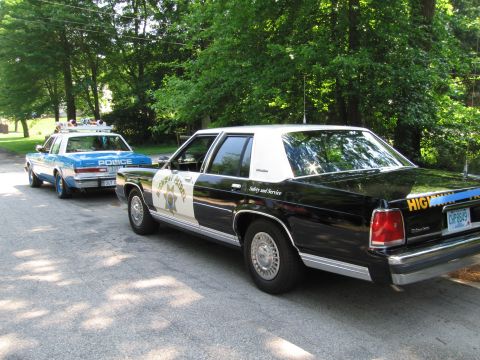 1991 Ford Crown Victoria - A black and white highway patrol car is parked behind a blue and white police car.