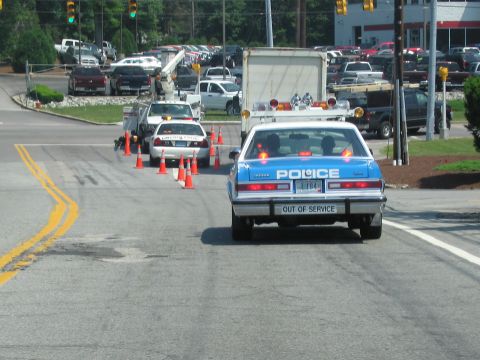 1991 Ford Crown Victoria - A police car marked 'OUT OF SERVICE' is behind traffic cones, with other vehicles in the background.