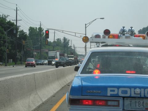 1991 Ford LTD Crown Victoria - Rear corner of a blue Dodge POLICE sedan with red-lit roof beacons waiting at a traffic light beside a highway barrier.