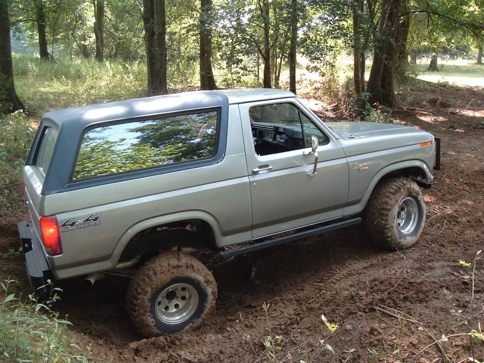 Lifted silver Ford Bronco with mud tires on a muddy trail in the woods, rear three-quarter view