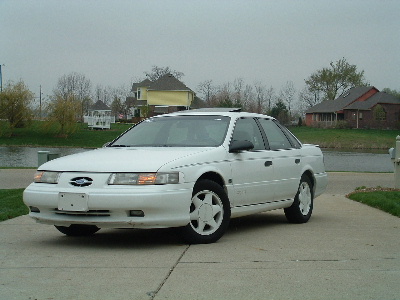 1993 Ford Taurus - A white 1993 Ford Taurus sedan is parked on a driveway in front of houses and trees.