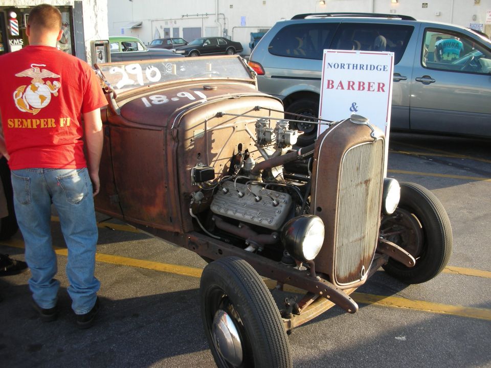 Rusty hot rod with exposed engine and a person in a red shirt standing next to it