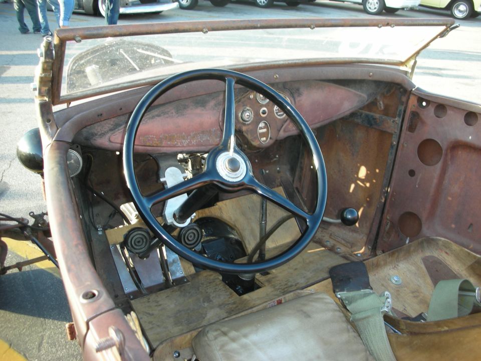 Steering wheel and dashboard of a rusty, old convertible. Floor is wood, seats are worn leather with a green seatbelt