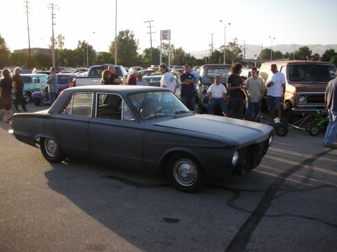 1998 Ford Crown Victoria - A group of people gather around a matte black sedan parked in a lot with other vehicles.